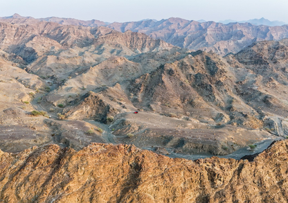 Panoramic aerial view of rugged UAE mountain ranges with a red 4x4 parked on a ridge.